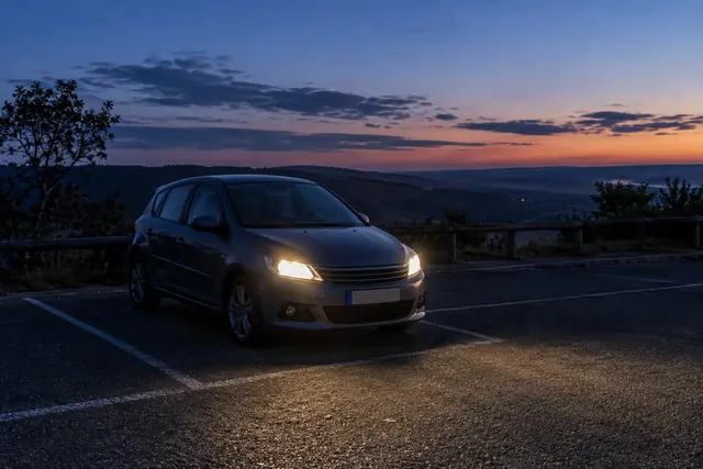 Illustration IA : Une voiture à l’arrêt dans une aire de repos au crépuscule, avec ses feux allumés.